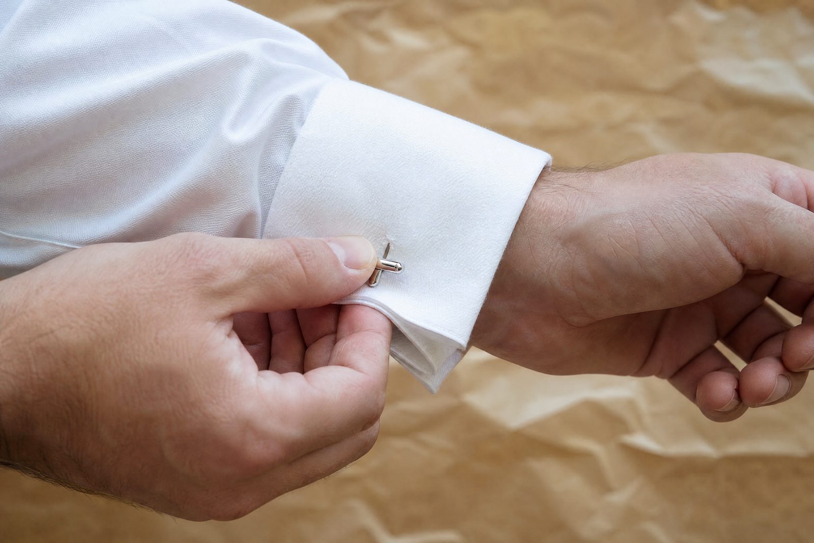 Two hands fastening a silver cufflink through the folded-back French cuff of a white shirt.